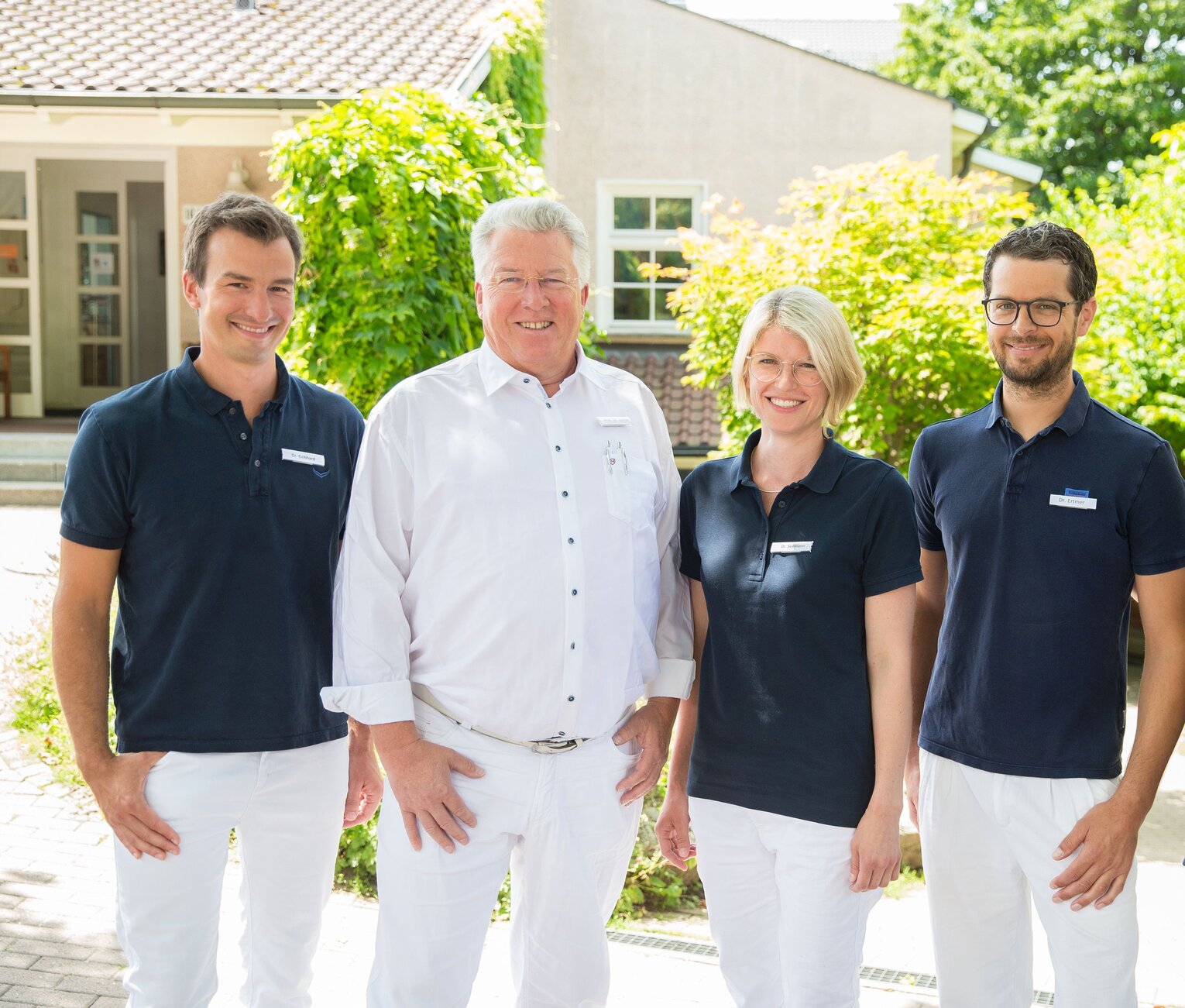 Ärzte der Orthopädie Tettnang Vier lächelnde Personen in Berufsbekleidung stehen vor einem Gebäude mit grünem Pflanzenwuchs.