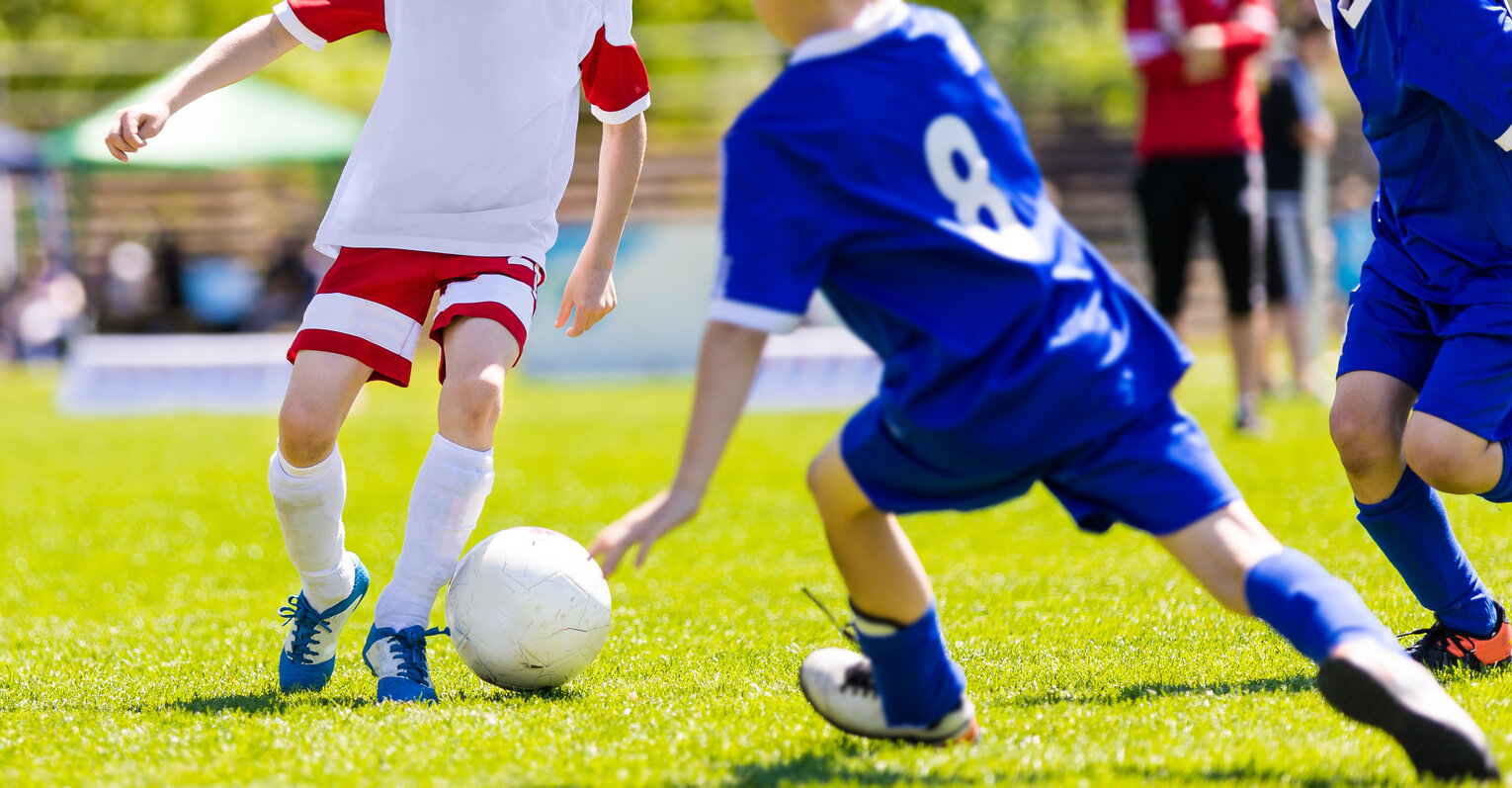 Kinder in roten und blauen Sportkleidungen spielen Fußball auf einem grünen Spielfeld.