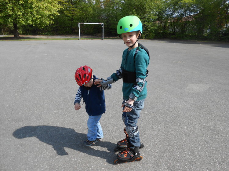 Ein älteres Kind mit Helm und Schutzausrüstung fährt Inlineskates und hält die Hand eines jüngeren Kindes mit rotem Helm auf einem leeren Sportplatz.