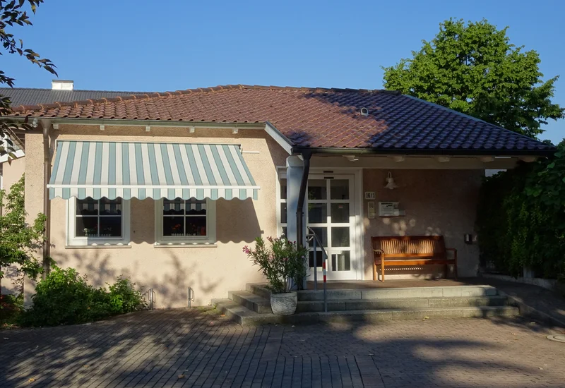 Orthopädie Tettnang von Außen Ein einzelnes beige Wohnhaus mit braunem Dach, markanter blau-weißer Stoff-Markise über zwei Fenstern und einer holzfarbenen Bank vor dem Hauseingang bei Sonnenschein.
